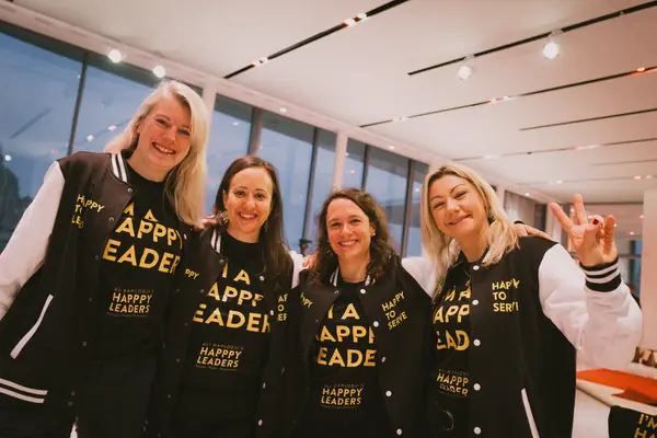 Vier lachende Frauen aus dem Team in schwarz-weißen College-Jacken „Happpy to serve“ und schwarzen Shirts „I’m a Happpy Leader“, eine zeigt das Victory-Zeichen