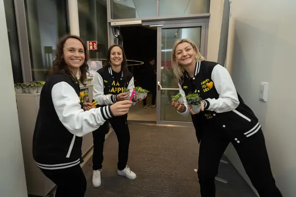 Drei Frauen in College-Jacken, eine mit der Aufschrift „Happy to serve“, lachen in die Kamera und halten kleine Topfpflanzen